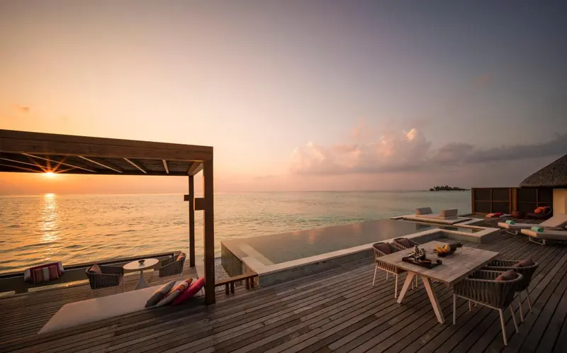 Terrasse en bois avec piscine et mobilier de détente face à la mer au coucher du soleil.