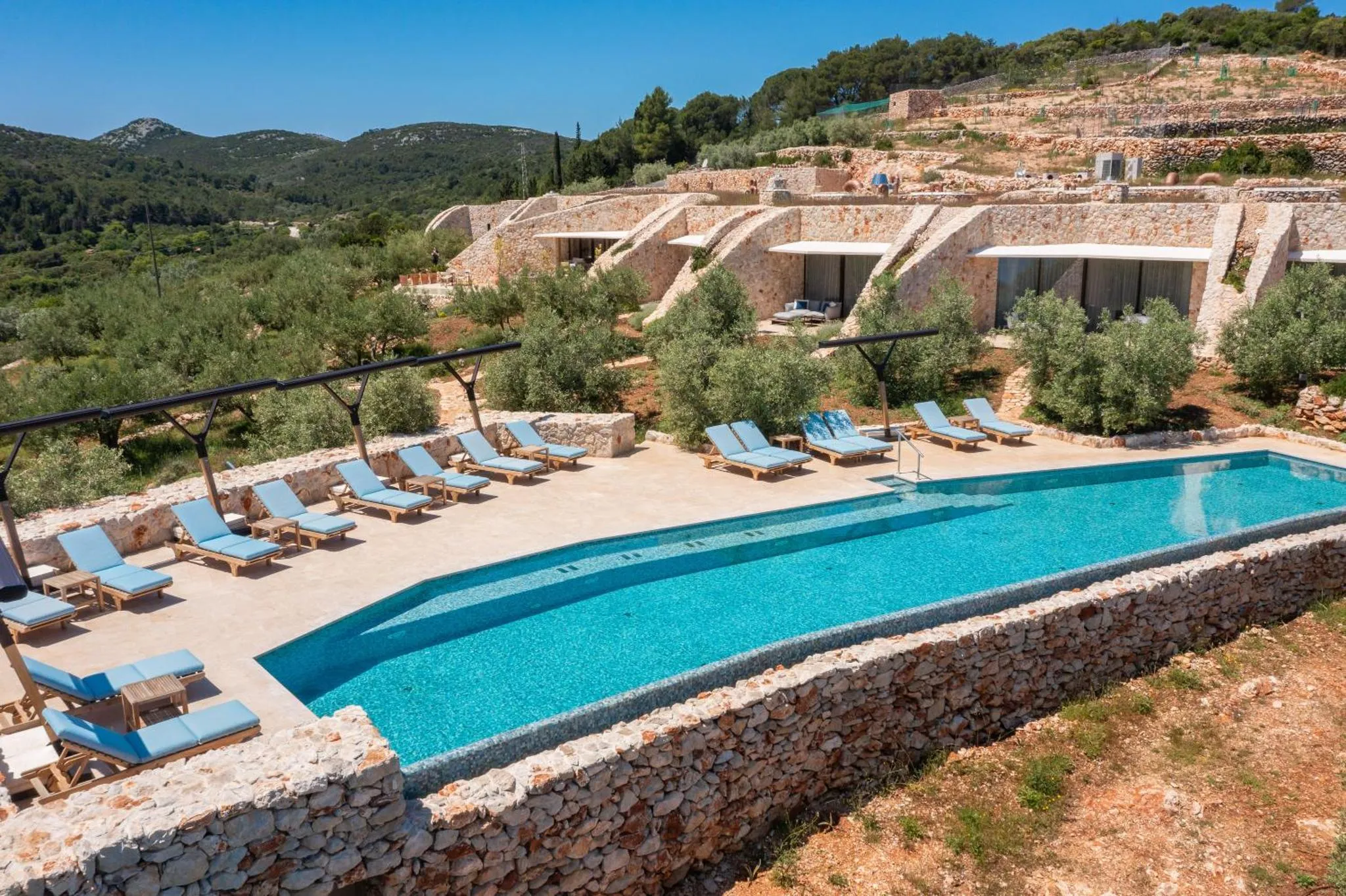 Piscine rectangulaire bordée de transats et d'une terrasse ombragée, avec des bâtiments en pierre en arrière-plan.