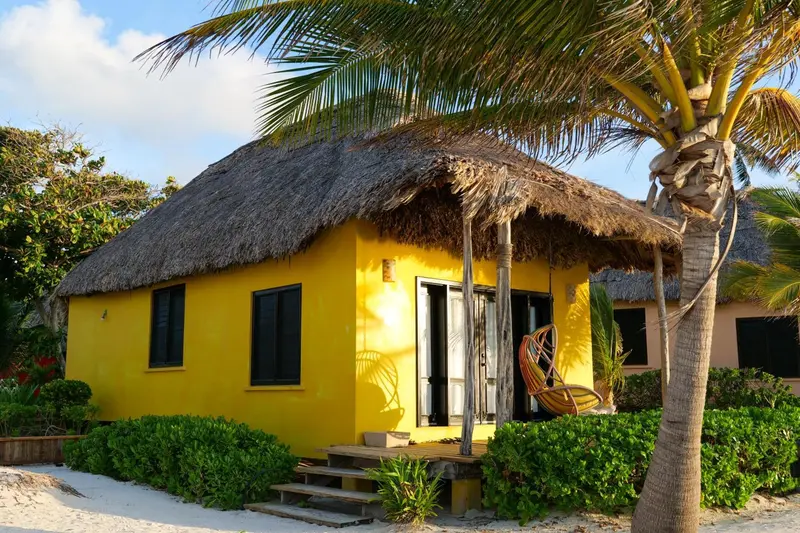 Une petite maison jaune aux toits de chaume se trouve sur une plage de sable blanc.