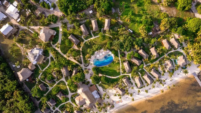 Vue aérienne d'un complexe de bungalows au toit de chaume entourés de végétation tropicale et bordant une plage.