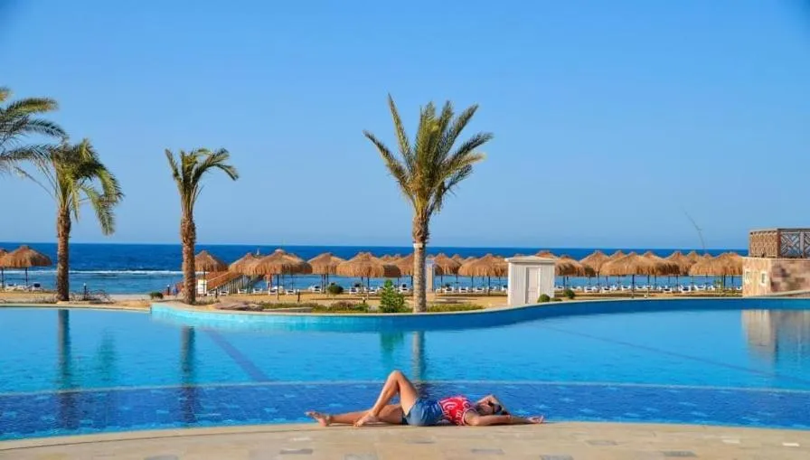 Une femme se relaxe au bord d'une piscine turquoise avec des palmiers en arrière-plan.