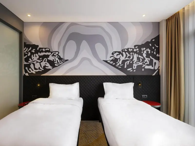Deux lits simples avec oreillers blancs dans une chambre décorée d'une fresque murale abstraite.