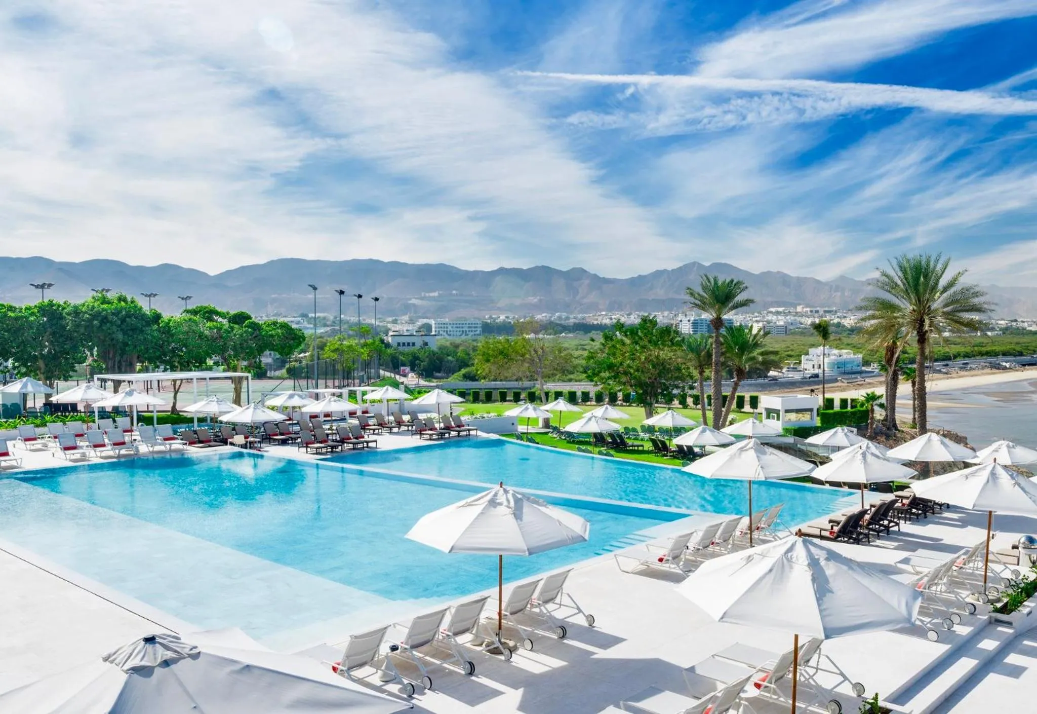 Une piscine turquoise bordée de transats blancs sous des parasols, avec une vue sur la mer et des montagnes.