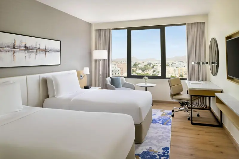 Chambre lumineuse avec deux lits et un espace de travail près d'une fenêtre.
