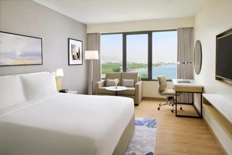 Chambre lumineuse avec un lit blanc, un coin salon et une vue sur la mer.