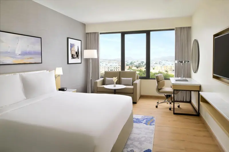 Une chambre lumineuse avec un lit, un coin salon et un bureau donnant sur une vue urbaine.