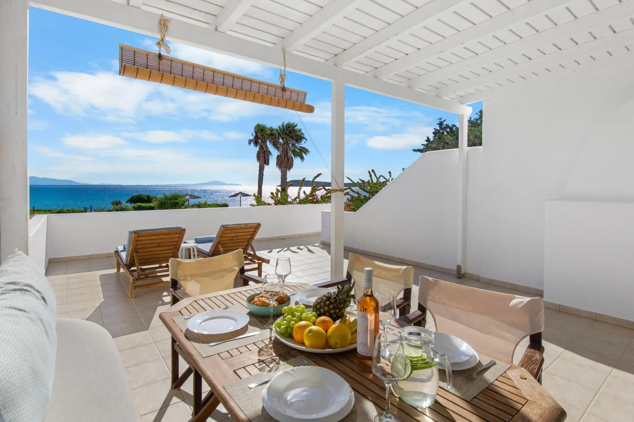 Une table dressée avec des fruits et des boissons sur une terrasse ensoleillée avec vue sur la mer.