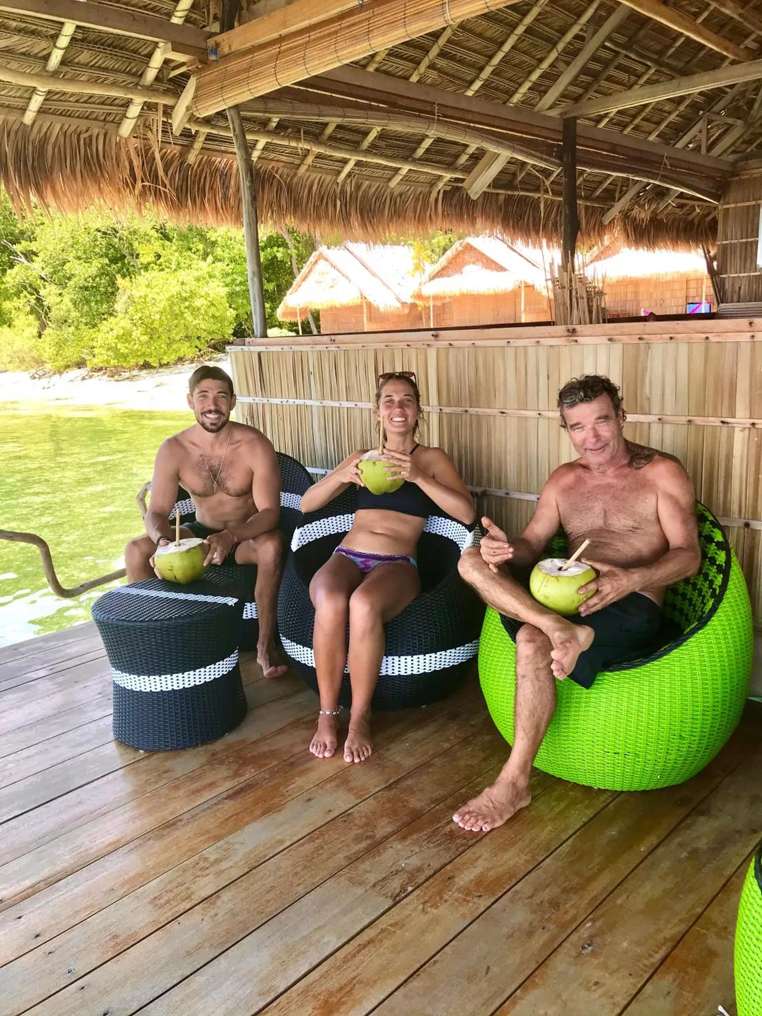 Trois personnes sourient en sirotant des boissons fraîches sur une terrasse en bois.