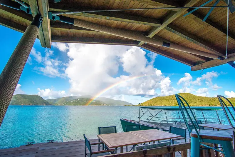 Terrasse ombragée donnant sur une baie turquoise et un arc-en-ciel.