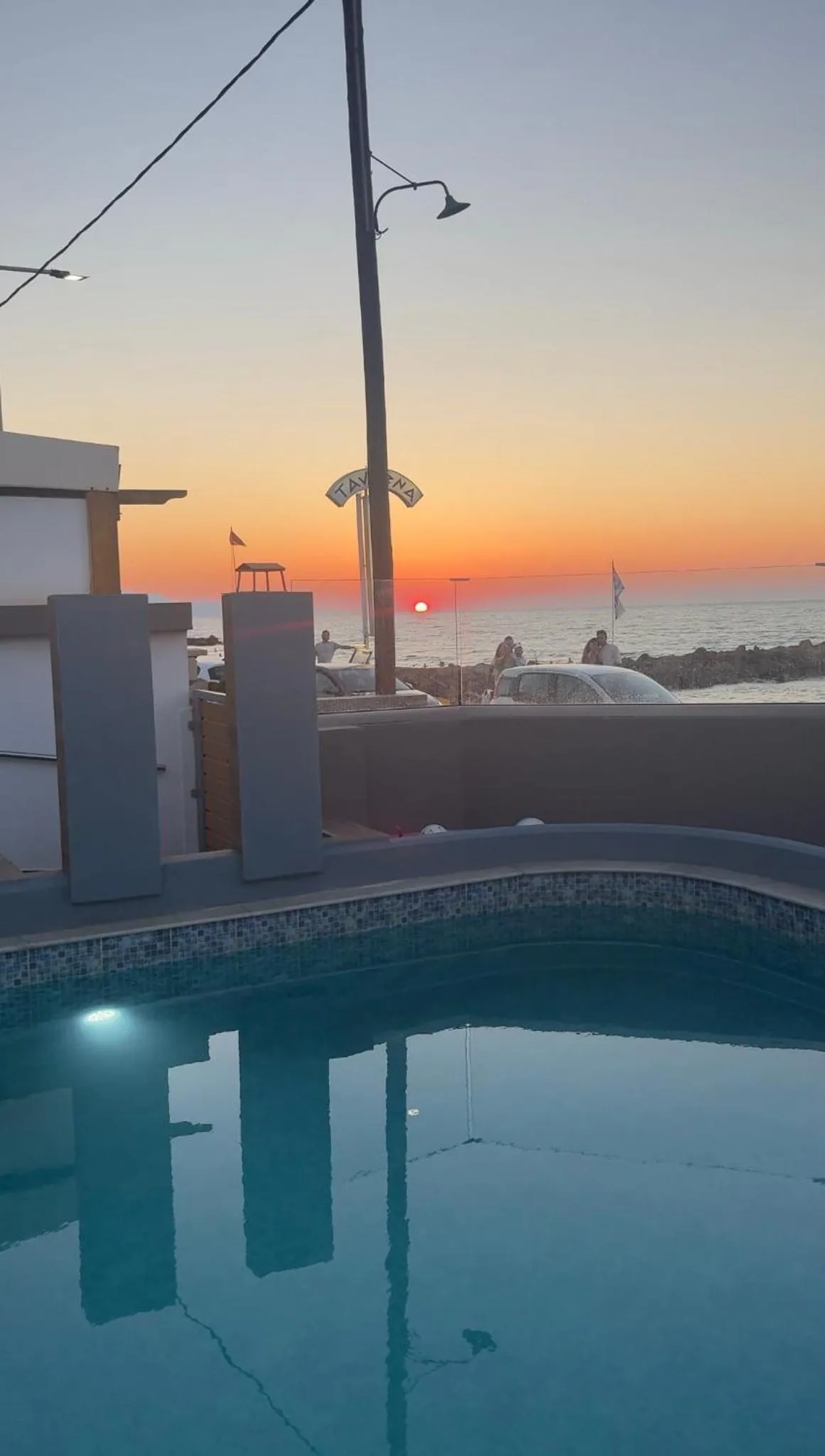 Une piscine au premier plan avec une vue sur la mer et un coucher de soleil orangé.