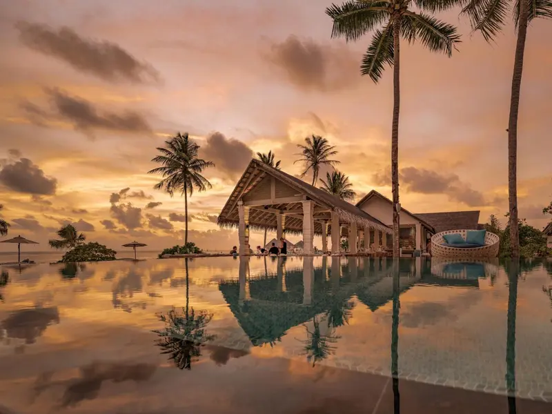 Une piscine à débordement reflète le ciel orangé au coucher du soleil, avec des palmiers et des structures balnéaires en arrière-plan.