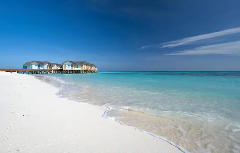 Plage de sable blanc bordée d'eau turquoise et de bungalows sur pilotis.