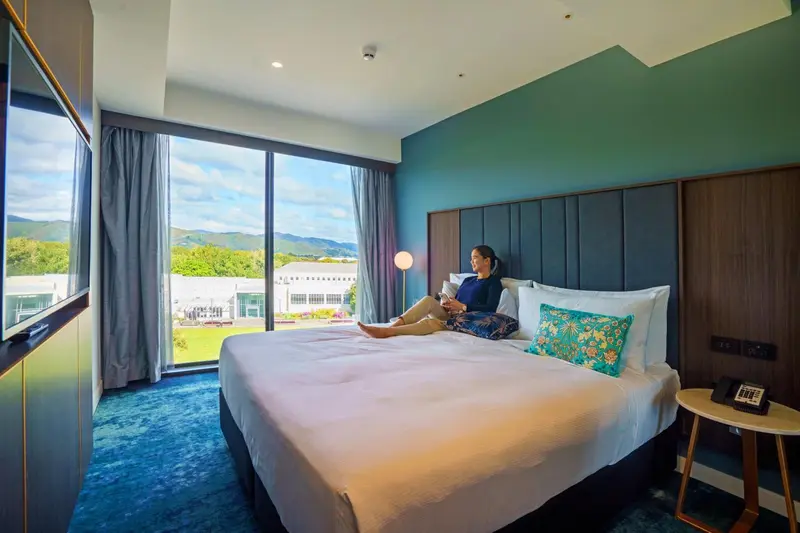 Une personne assise sur un lit dans une chambre lumineuse avec vue sur un paysage verdoyant.
