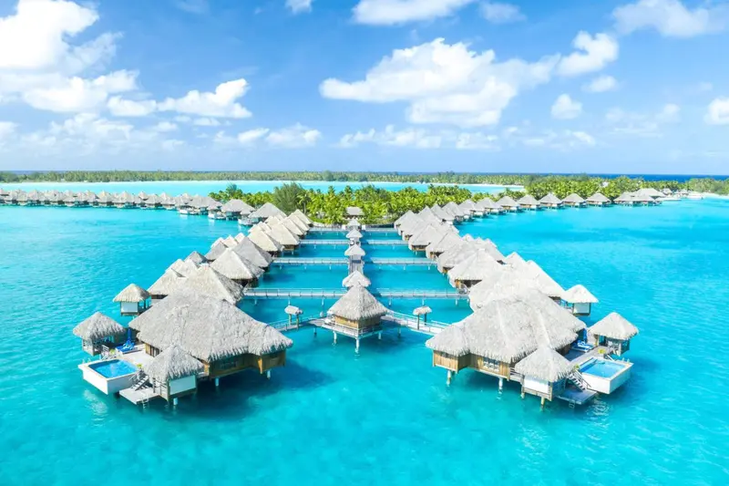 Vue aérienne d'un ensemble de bungalows sur pilotis au-dessus d'une eau turquoise.