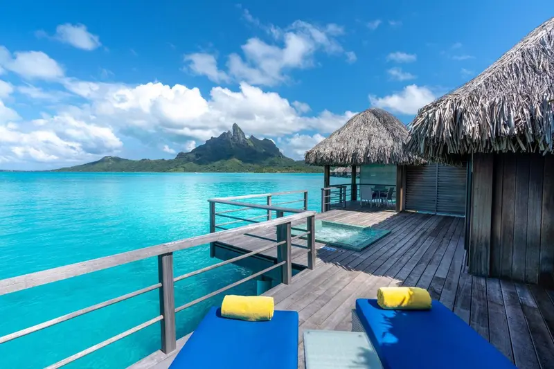 Terrasse en bois avec transats donnant sur une eau turquoise et une montagne au loin.