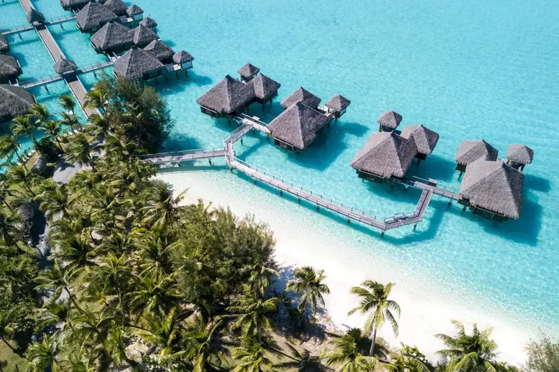 Vue aérienne de bungalows sur pilotis au-dessus d'une eau turquoise et d'une plage de sable blanc.