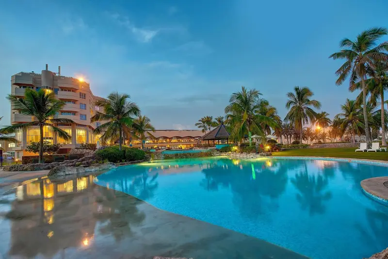 Une piscine turquoise au premier plan, avec un bâtiment illuminé et des palmiers en arrière-plan.
