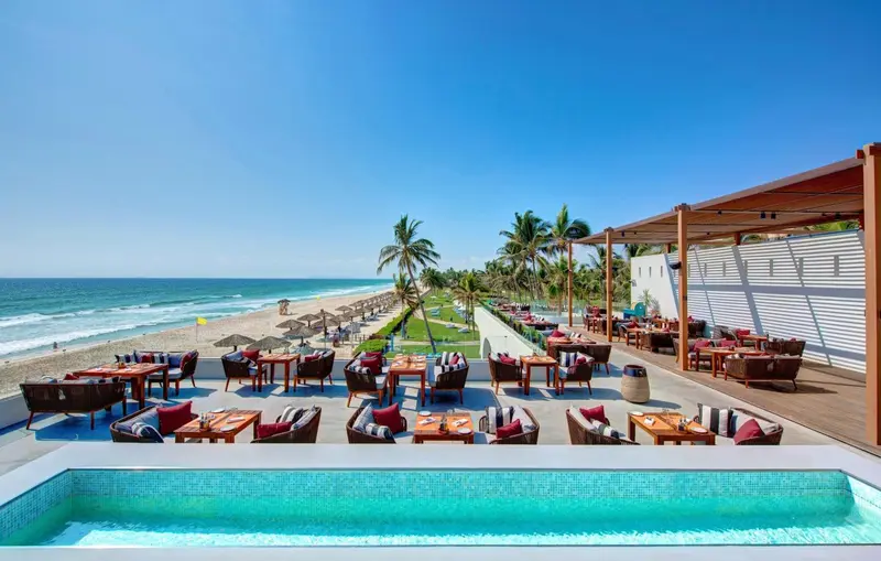 Terrasse avec tables et chaises donnant sur une plage de sable fin et une mer bleue.
