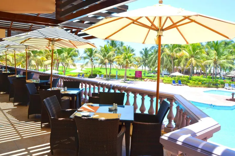 Terrasse ombragée avec tables et chaises donnant sur une piscine et la mer.