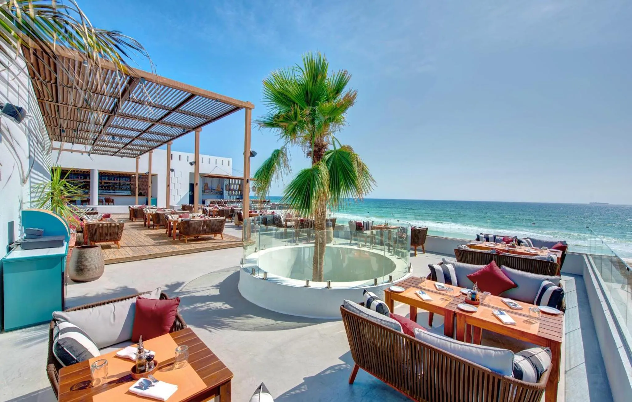 Terrasse ombragée avec tables et canapés donnant sur la mer.
