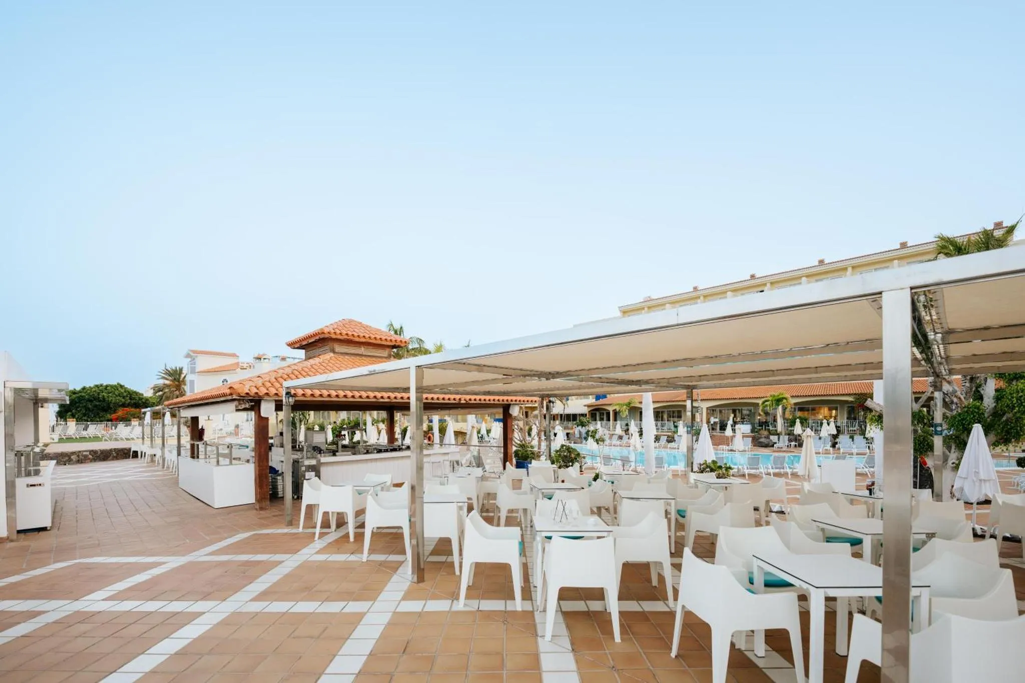 Terrasse ombragée avec tables et chaises blanches près d'une piscine.