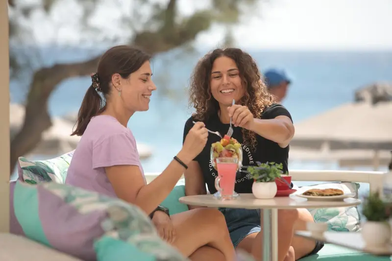 Deux femmes sourient en dégustant un dessert sur une terrasse ensoleillée.