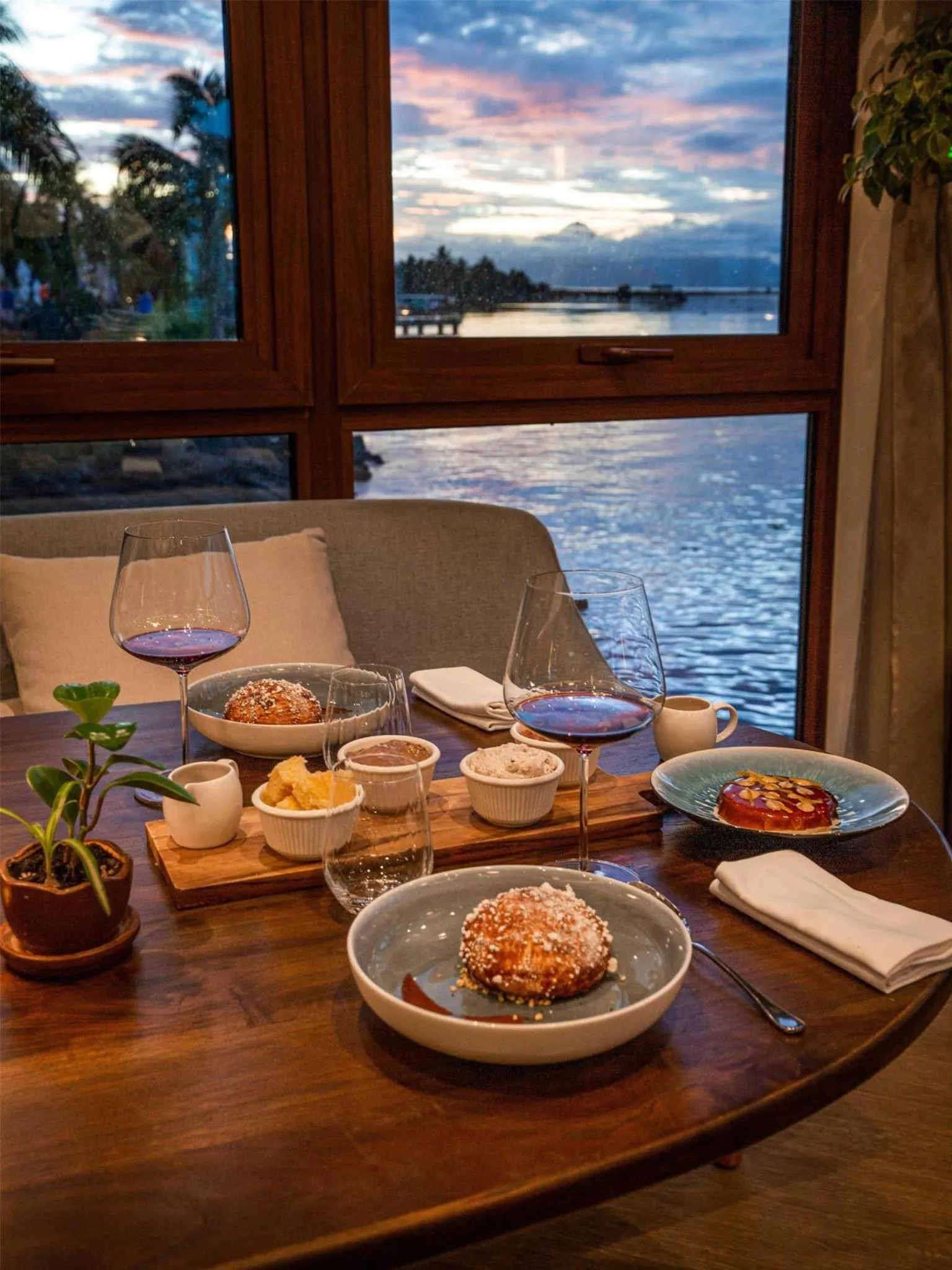 Une table ronde en bois présente un repas avec des plats et des verres de vin, devant une grande fenêtre offrant une vue sur la mer et un coucher de soleil.