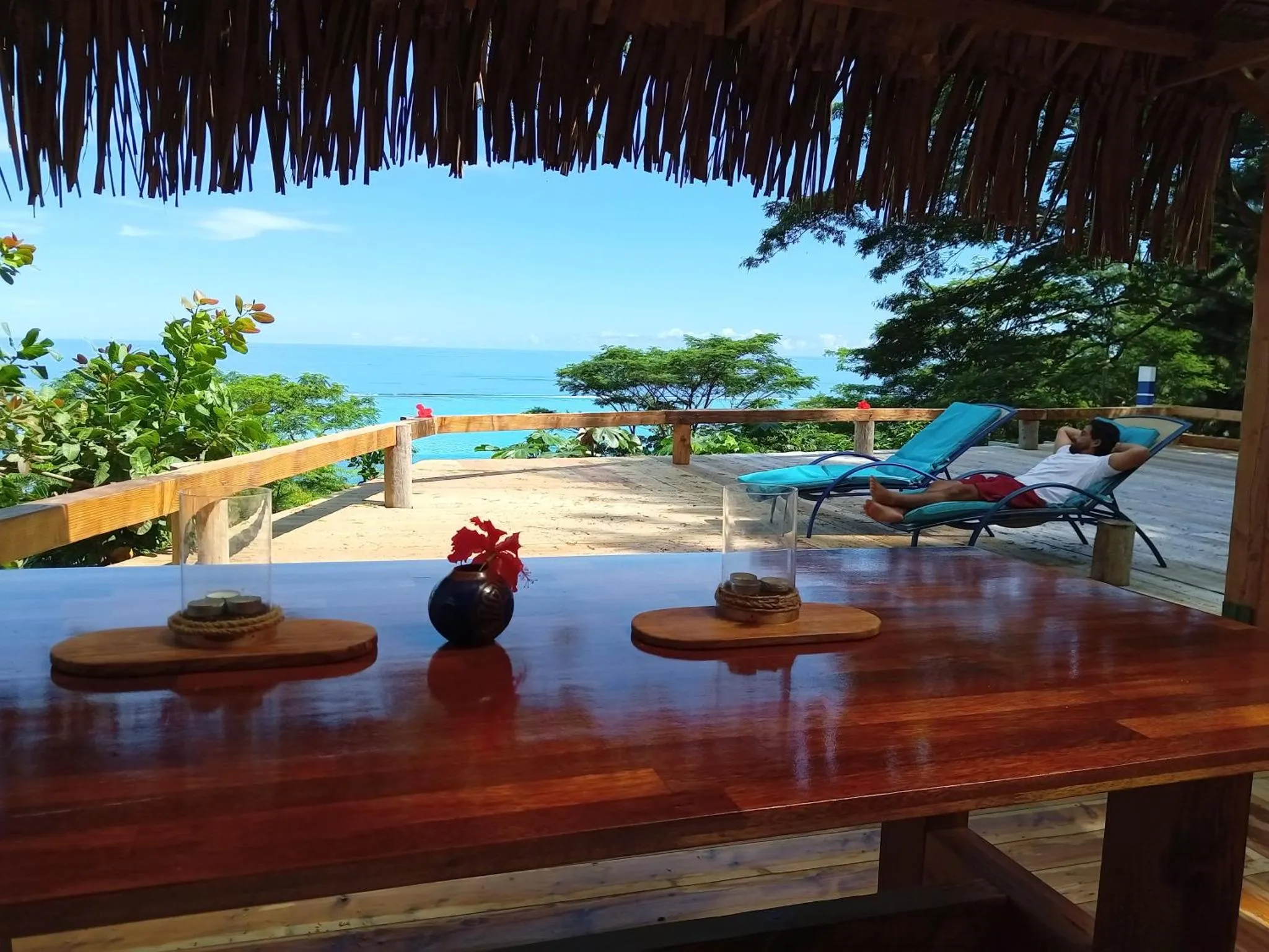 Une table en bois est mise en scène sur une terrasse ombragée avec vue sur la mer.