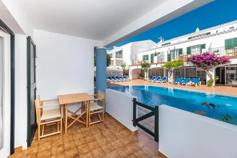 Terrasse avec table et chaises donnant sur une piscine et des bâtiments blancs.