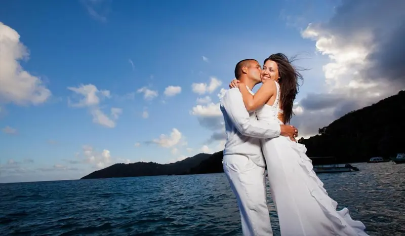 Un couple s'embrasse sur une plage ensoleillée avec une mer turquoise en arrière-plan.