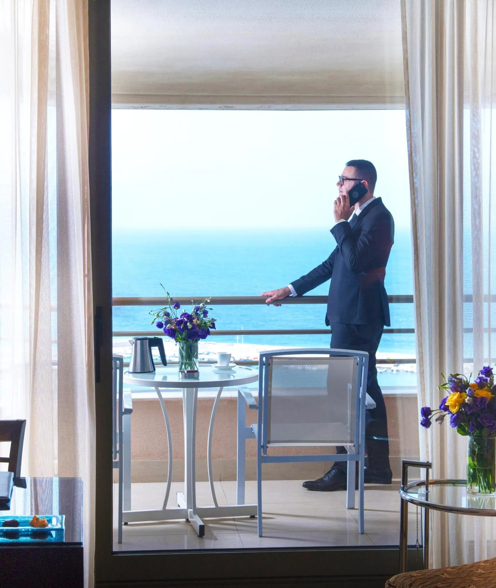 Un homme en costume parle au téléphone sur une terrasse avec vue sur la mer.
