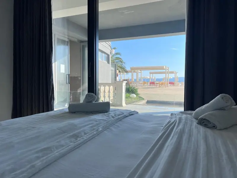 Un lit blanc est visible à travers une baie vitrée donnant sur une plage.