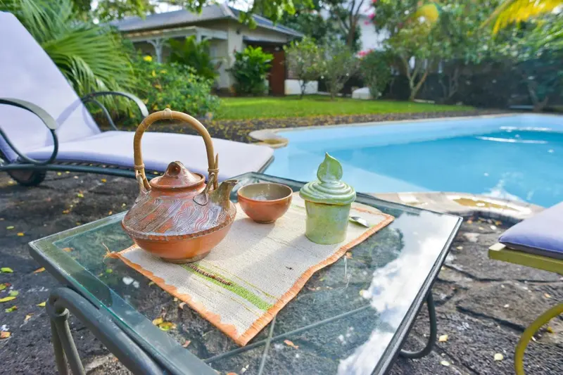 Une table basse en verre présente une théière et des tasses sur une terrasse ensoleillée près d'une piscine.