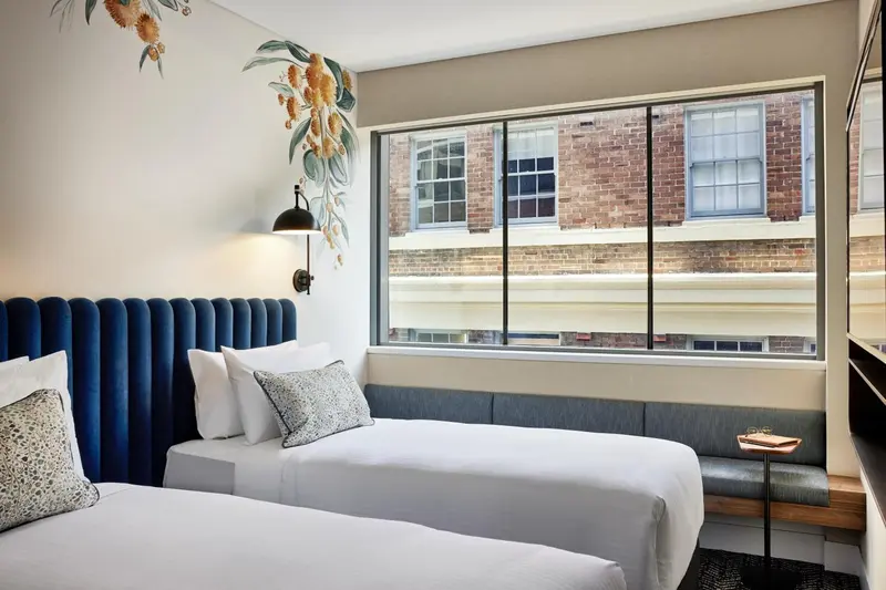 Une chambre avec deux lits simples et une grande fenêtre donnant sur un bâtiment en briques.