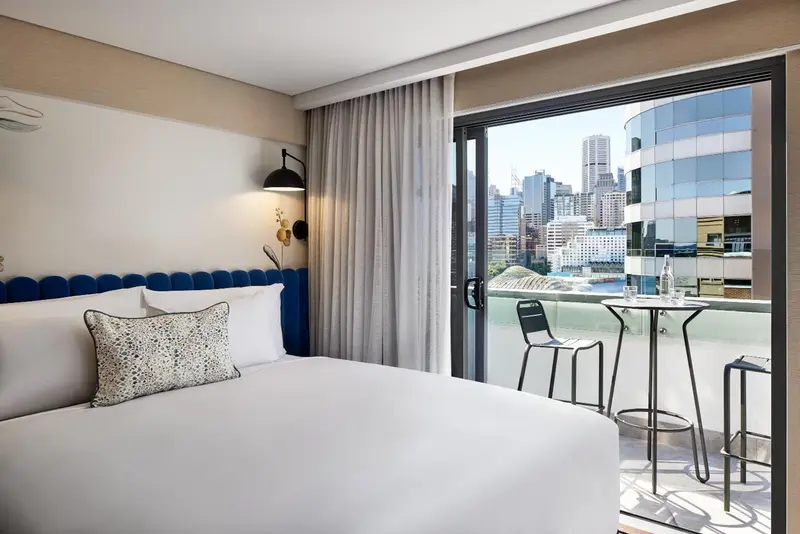 Chambre lumineuse avec un grand lit et un balcon offrant une vue sur la ville.