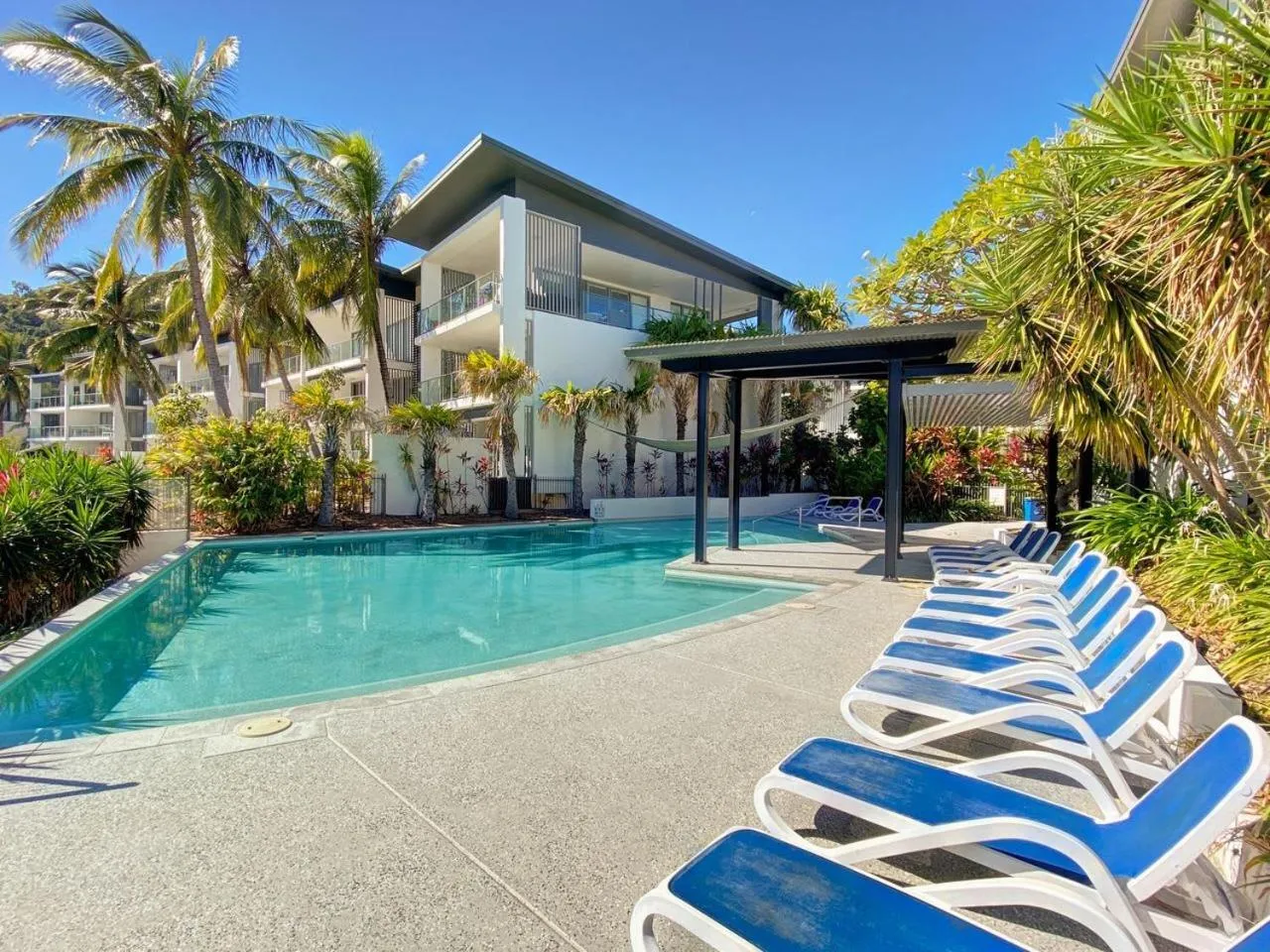 Piscine turquoise entourée de transats bleus et de végétation tropicale devant un bâtiment moderne.