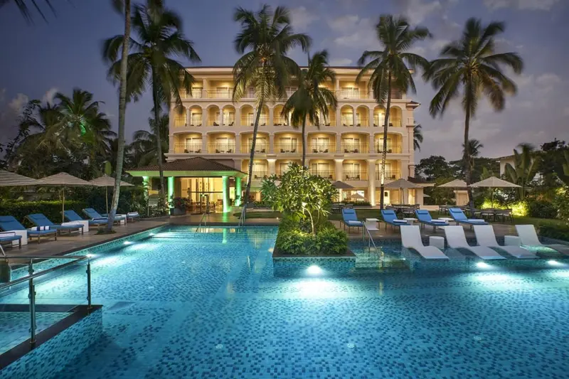 Bâtiment illuminé au milieu d'une végétation tropicale, avec une piscine au premier plan.