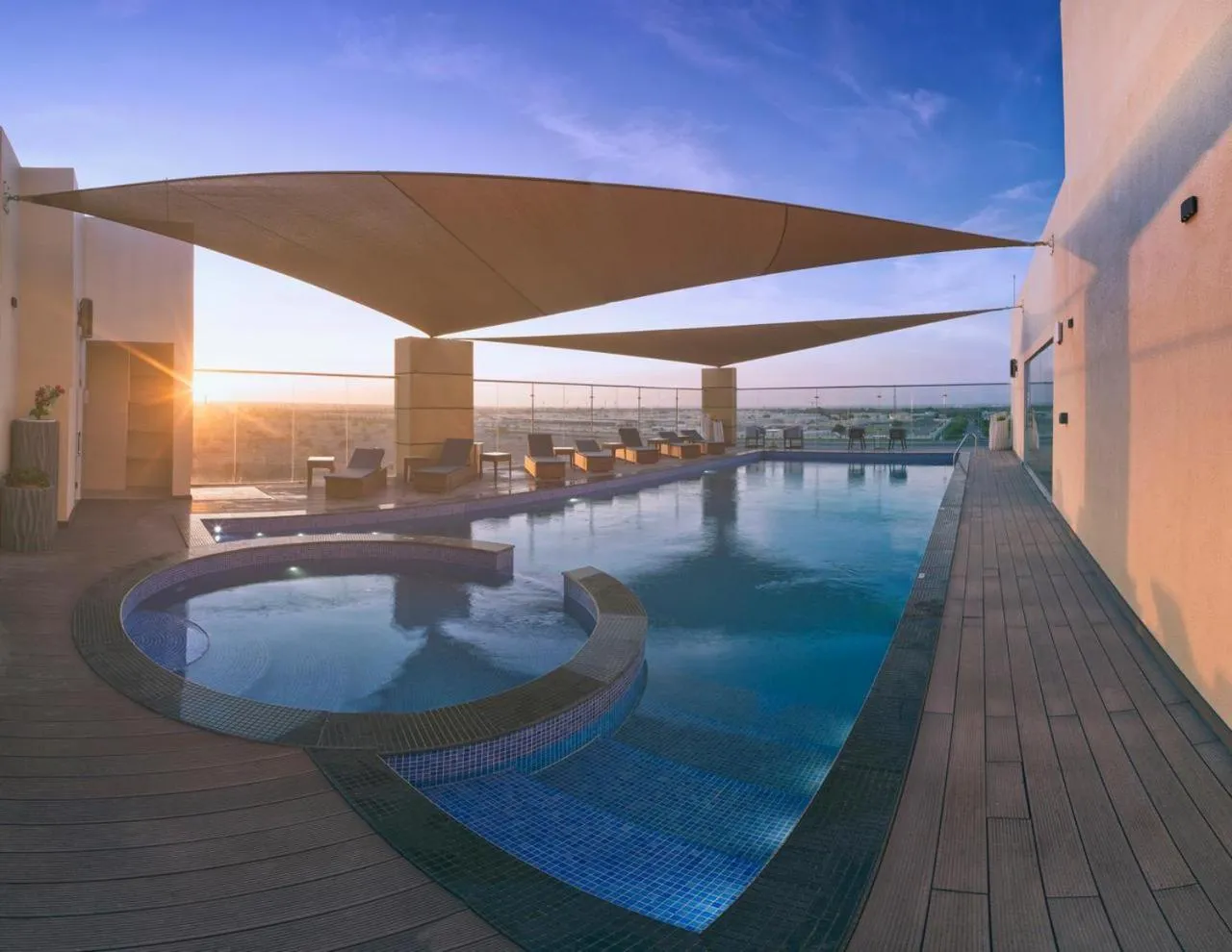 Une piscine rectangulaire avec un jacuzzi intégré, entourée d'une terrasse en bois et ombragée par des voiles.