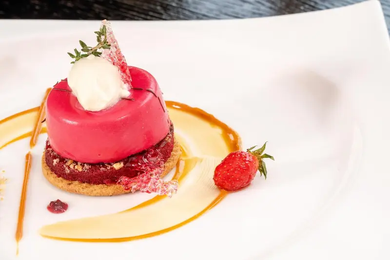 Un dessert rouge vif est présenté sur une assiette blanche avec une sauce dorée et une fraise.