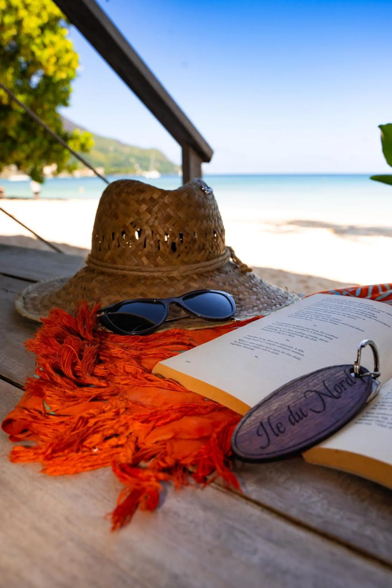 Un chapeau de paille, des lunettes de soleil et un livre reposent sur une table en bois face à la mer.