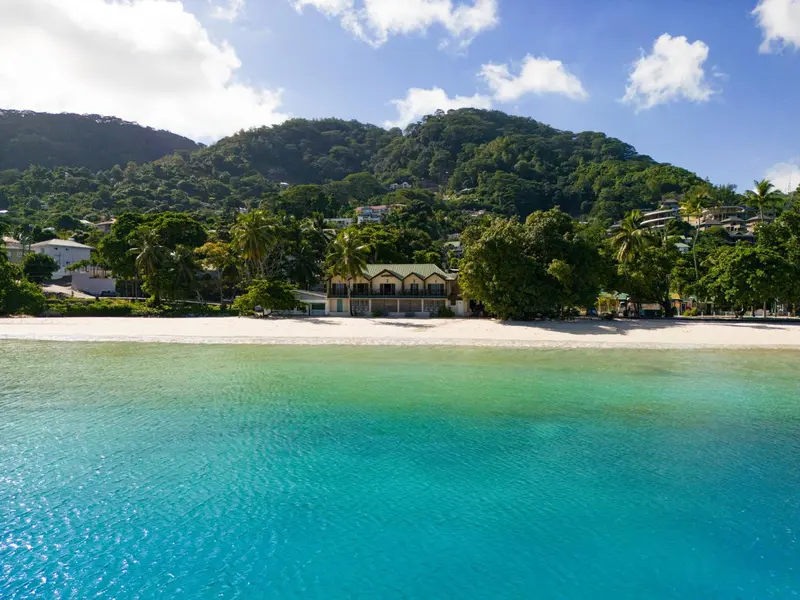Vue aérienne d'une plage de sable blanc bordée de végétation luxuriante et de bâtiments clairs.