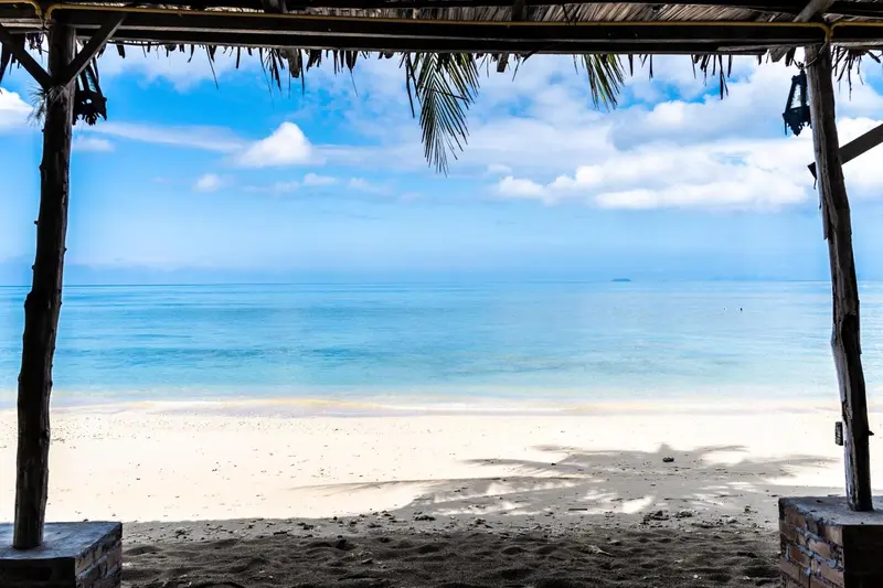 Vue sur une plage de sable blanc et une mer turquoise à travers une structure en bois et palmes.