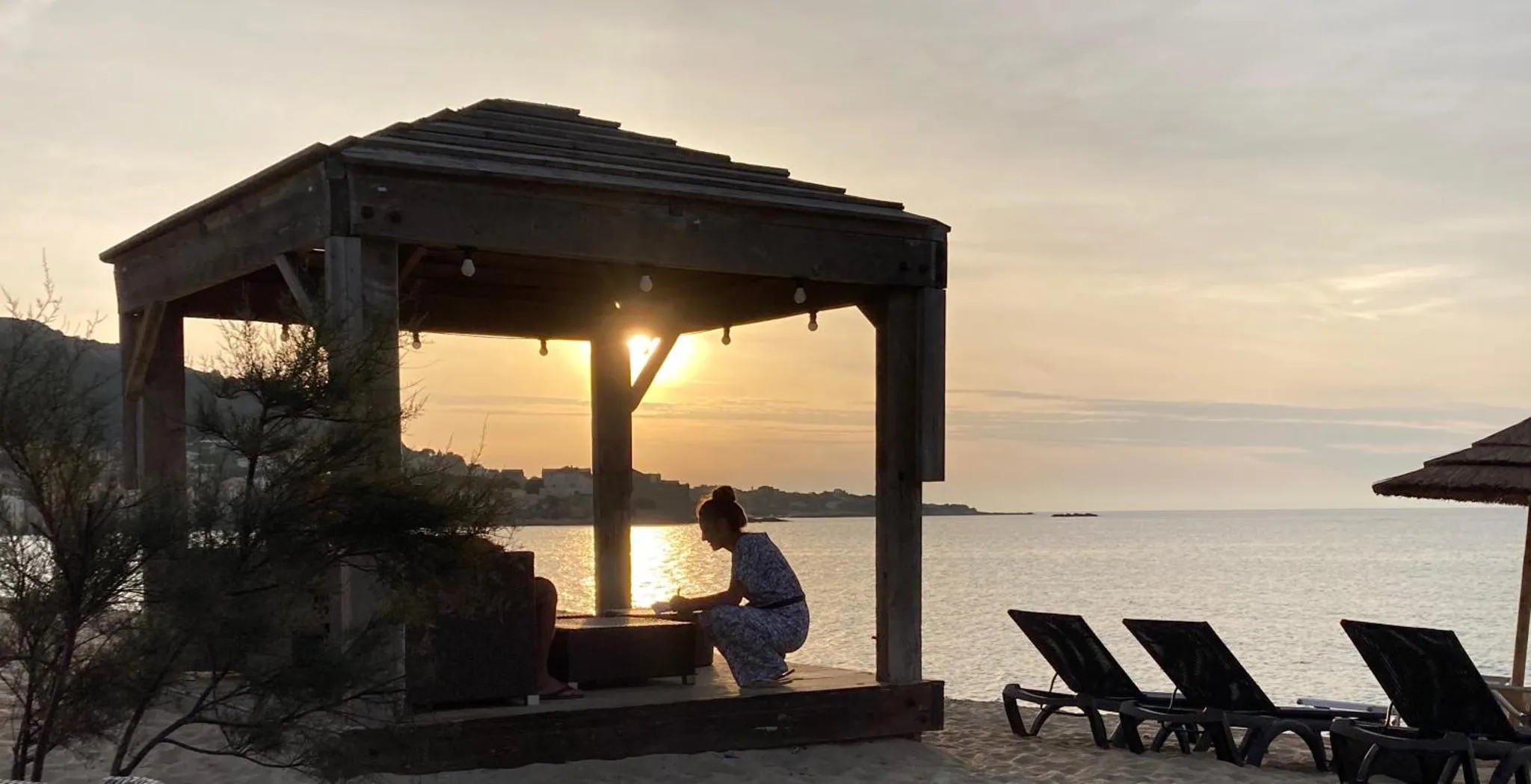 Une personne est assise sous un abri en bois face à la mer au coucher du soleil.