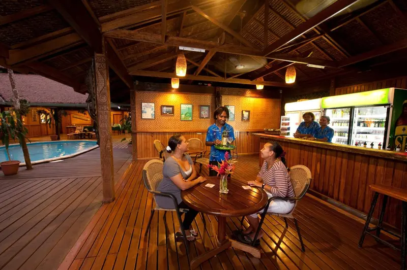 Des clients sont servis au comptoir d'un bar en bois, près d'une piscine.