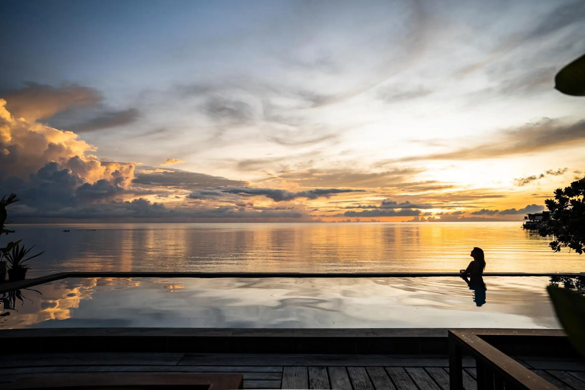 Une personne se tient au bord d'une piscine à débordement face à un coucher de soleil sur l'océan.