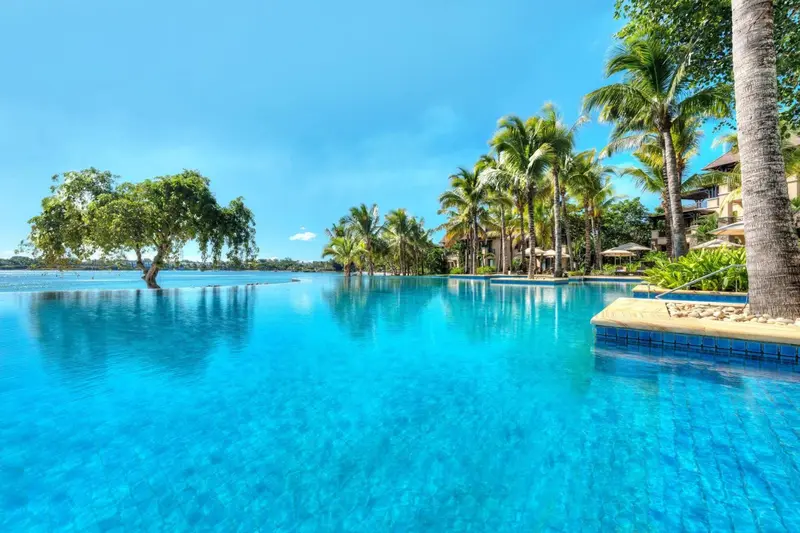 Une piscine à débordement turquoise reflète le ciel bleu et les palmiers environnants.
