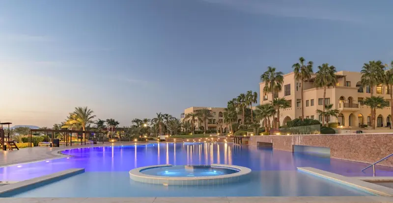 Une piscine illuminée au crépuscule devant un bâtiment beige.