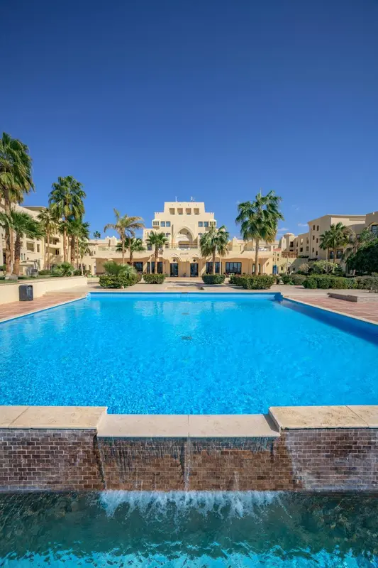 Vue d'une piscine turquoise devant un bâtiment beige et blanc sous un ciel bleu.