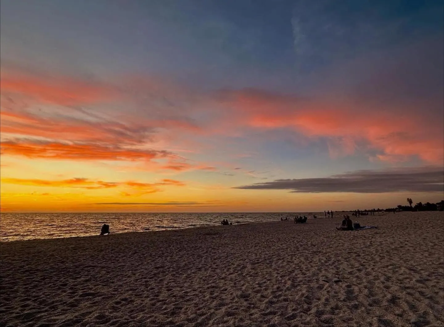Coucher de soleil sur une plage de sable avec quelques personnes assises ou allongées.
