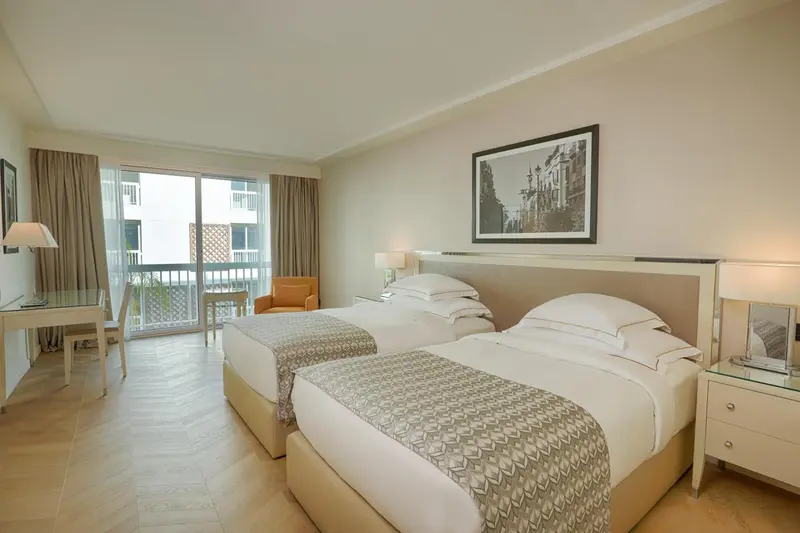 Une chambre lumineuse avec deux lits simples et un fauteuil près d'une baie vitrée.
